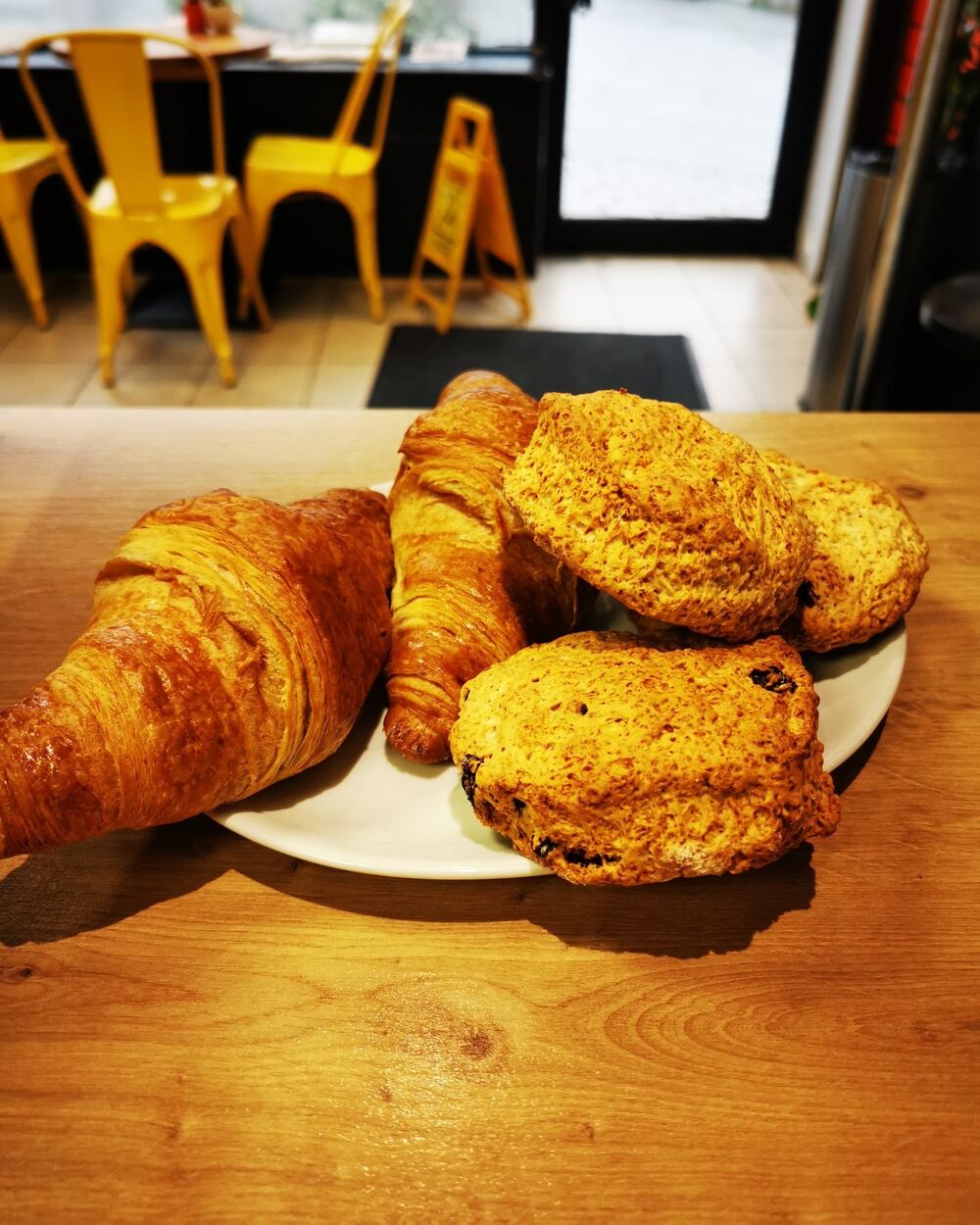 Croissants and scones on a plate on a wooden counter, with yellow metal chairs behind